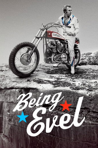 The black-and-white image features a rugged motorcyclist in a white and blue racing suit, poised on a dirt bike against a rugged, rocky backdrop. The poster's bold, stylized text "Being Evel" with star accents conveys a bold, adventurous tone. The mood is one of rugged freedom and determination.