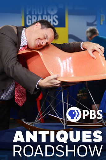 A man in a suit and tie leans over a vibrant orange chair, appearing to be in distress or exhaustion. The PBS logo and "Antiques Roadshow" text are prominently displayed at the bottom. The scene captures a humorous and unexpected moment, blending humor with the show's quirky atmosphere.