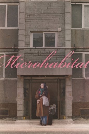 The poster features a solitary figure in a vintage coat and hat standing before a weathered brick building, evoking a nostalgic, slightly melancholic atmosphere. The muted color palette of browns and grays enhances the sense of timelessness and solitude. The title "Microhabitat" is elegantly writ...
