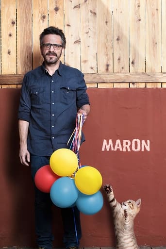 A man in a dark shirt stands beside a colorful balloon cluster, with a cat reaching up to touch the yellow balloon. The poster features a warm, inviting mood against a rustic wooden backdrop and a red wall with "MARON" written in bold white letters.