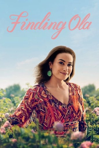 The poster features a woman with long brown hair, wearing a vibrant red and orange patterned blouse, standing amidst lush greenery and pink flowers. The soft, dreamy lighting enhances the romantic and nostalgic mood of the scene.