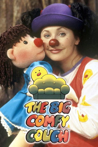 The poster features a woman in a purple hat and red nose, holding a plush doll with a yellow duck on its head. The background is warm and cozy, with soft lighting. The title "The Big Comfy Couch" is displayed in colorful, playful letters. The overall mood is cheerful and inviting, suggesting a re...