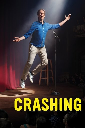 A man in a blue shirt and beige pants leaps joyfully on a stool, arms outstretched against a spotlight. The poster features bold yellow text at the bottom, evoking a sense of excitement and humor.