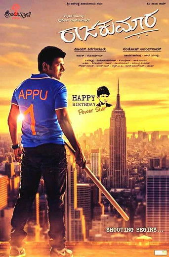 The poster features a man in a blue shirt with "APPU" printed on it, standing against a vibrant sunset backdrop. The city skyline is illuminated by warm hues, creating a dramatic and energetic atmosphere. The title "Raajakumara" is prominently displayed in bold, stylized text, suggesting a blend ...