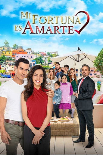 The poster features a vibrant scene with a man in a white shirt and a woman in a red top standing prominently in the foreground, surrounded by a diverse group of family members. The background showcases a picturesque Mediterranean-style town with colorful buildings and a clear blue sky. The warm ...