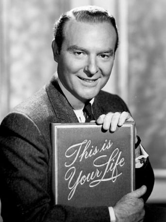 A smiling man in a suit holds a framed poster reading "This Is Your Life" against a softly lit background. The warm tones and confident pose convey a nostalgic, celebratory mood.
