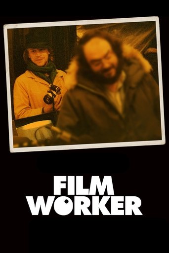The poster features a warm, sepia-toned photograph of two film workers in winter gear, one holding a camera and the other adjusting their fur-lined coat. Below the image, bold white text reads "FILM WORKER" against a stark black background, evoking a gritty, authentic atmosphere.