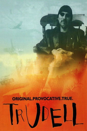 The poster features a man in a dark outfit sitting in a chair against a fiery orange and yellow background. The title "TRUDELL" is prominently displayed in bold, black letters at the bottom. The overall mood is intense and dramatic, suggesting a thriller or action movie.