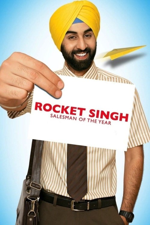 A smiling Sikh man in a yellow turban holds up a white card with "ROCKET SINGH SALESMAN OF THE YEAR" in bold red letters. The vibrant yellow and red colors against a blue background create a cheerful and energetic mood.