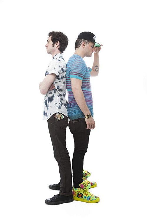 Two men stand back-to-back against a stark white background, exuding a rebellious and playful mood. One wears a floral shirt and black pants, while the other sports a striped shirt and bright green shoes. Their dynamic poses suggest camaraderie and humor, capturing the essence of the movie's ligh...