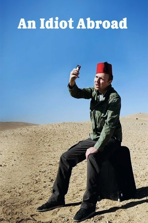The poster features a man in traditional attire sitting on a sand dune, holding a smartphone aloft. He wears a red fez and green jacket against a backdrop of vast desert under a clear blue sky. The title "An Idiot Abroad" is prominently displayed at the top, suggesting humor and cultural contrast...