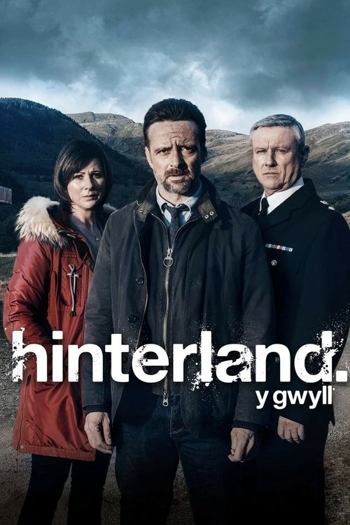 The poster features three characters standing against a dramatic, moody sky with rugged mountains in the background. The central figure wears a dark jacket, while the woman on the left dons a red coat with fur trim, and the man on the right sports a dark suit. The title "Hinterland" is prominentl...