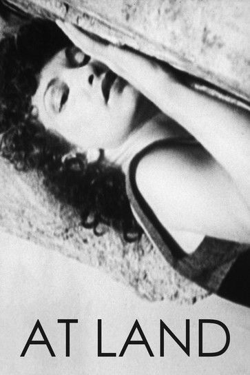 The black-and-white poster features a woman with dark, curly hair resting her head on a textured stone wall, her hand raised near her face. The mood is somber and reflective, evoking a sense of solitude and introspection. The title "AT LAND" is prominently displayed in bold, uppercase letters at ...
