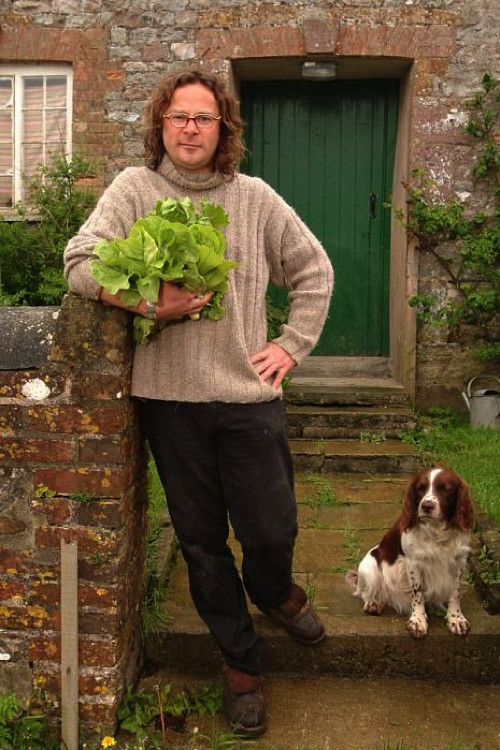 A man with glasses and a sweater holds a leafy plant, leaning against a rustic brick wall. Beside him sits a fluffy dog, and a green door stands in the background. The scene exudes a cozy, pastoral mood with earthy tones and a sense of tranquility.