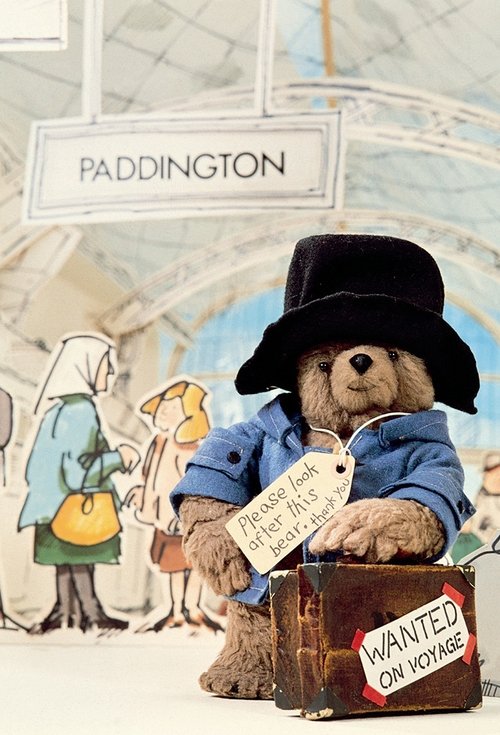 Paddington Bear, dressed in a blue coat and black hat, sits proudly beside a vintage suitcase labeled "WANTED ON VOLUME." Behind him, two children—a boy in a blue dress and a girl in a green dress—walk past, suggesting a lively, nostalgic atmosphere. The poster features a classic map background w...