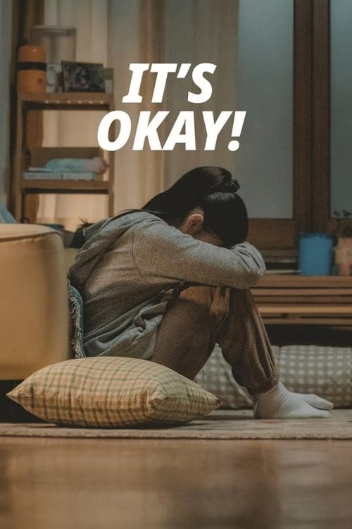 The poster features a woman sitting alone on a wooden floor, hugging her knees in despair. The warm, cozy room with soft lighting and a comfortable couch creates a somber mood. The bold white text "IT'S OKAY!" stands out against the muted tones, emphasizing the message of acceptance and healing.