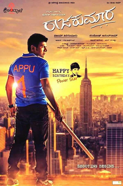 The poster features a man in a blue shirt with "APPU" printed on it, standing against a vibrant sunset backdrop. The city skyline is illuminated by warm hues, creating a dramatic and energetic atmosphere. The title "Raajakumara" is prominently displayed in bold, stylized text, suggesting a blend ...