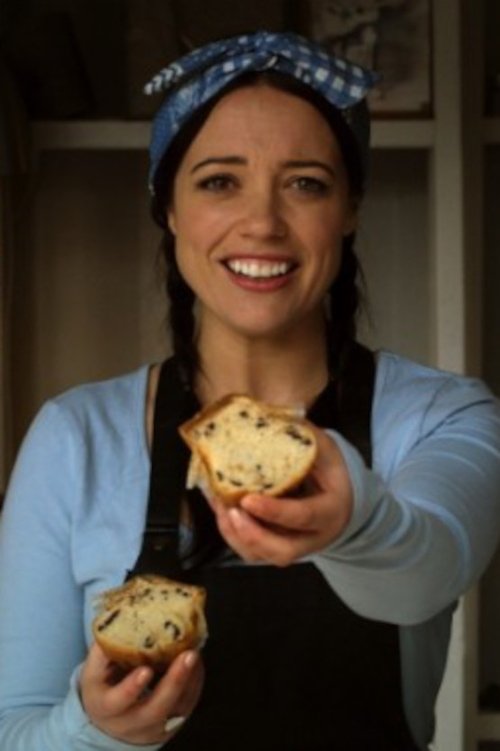 A woman in a blue shirt and black apron holds up a slice of bread with chocolate chips, smiling warmly against a softly lit kitchen backdrop. The warm tones and cheerful expression convey a sense of joy and nostalgia.